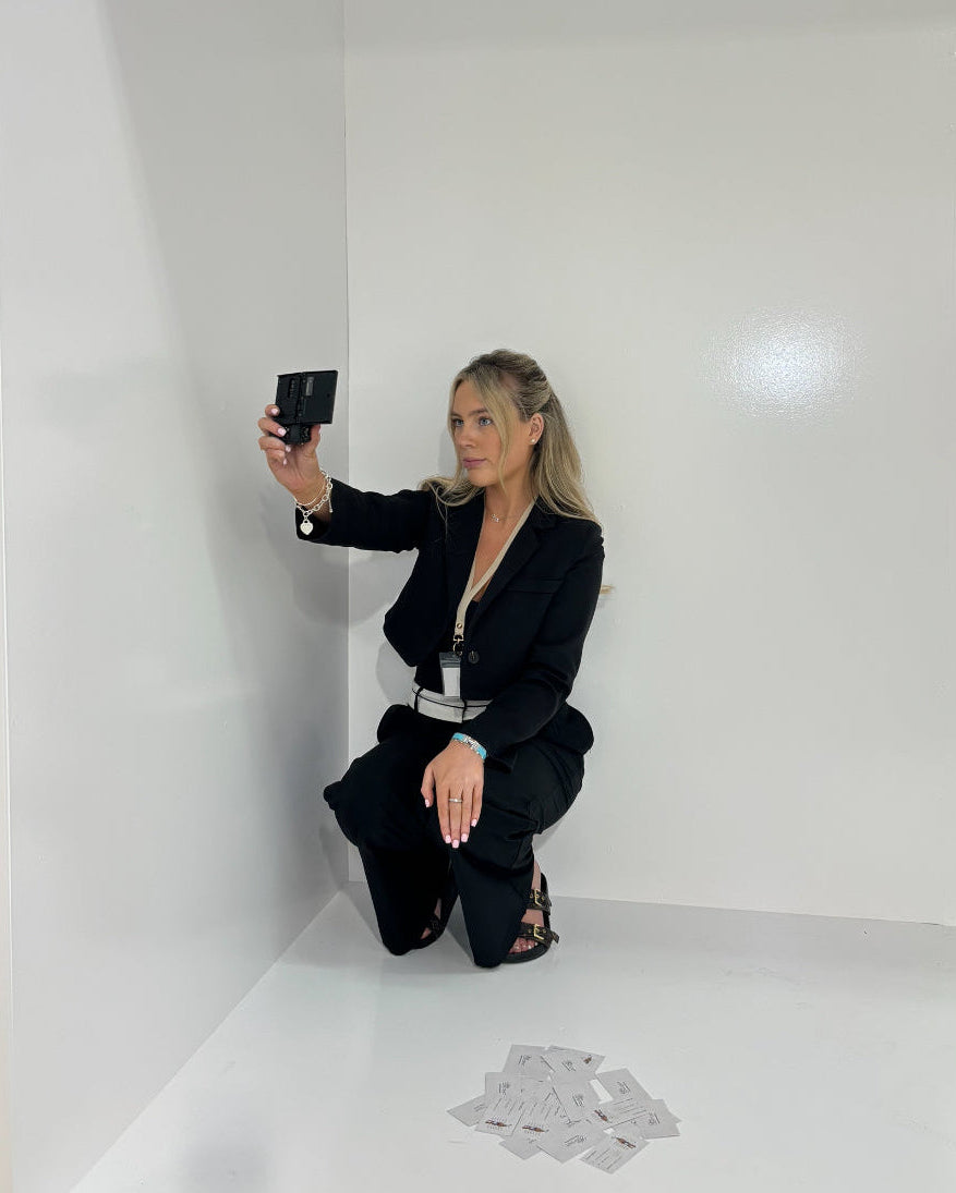Person in a black outfit taking a mirror selfie in a minimal white room with scattered cards on the floor.